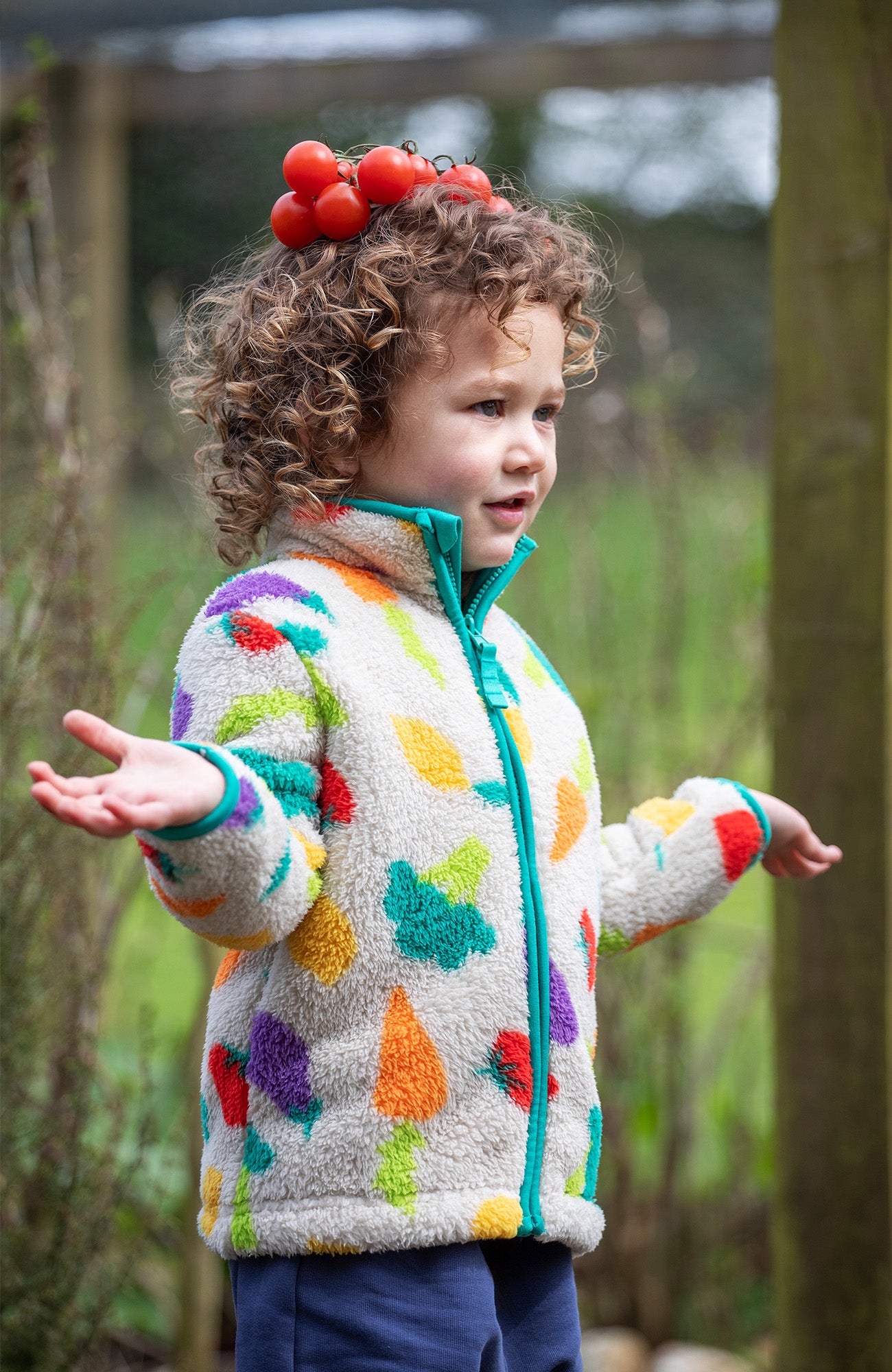 Child with curly brownish-blonde hair playfully balancing tomatoes on their head, outdoors in a garden with greenery in the background, wearing a cosy creamy white Frugi Ted Fleece featuring a fruit and vegetable print from the new Spring '25 range at Babipur.