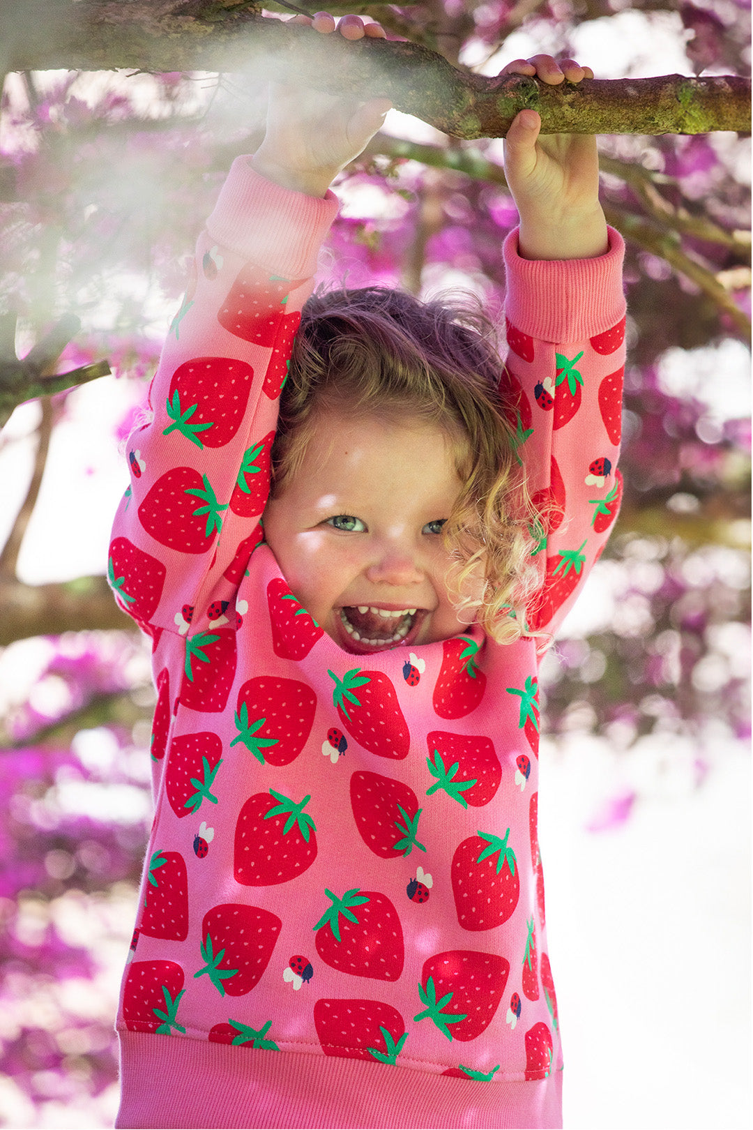 "A cheerful young child with curly blonde hair playfully hangs from a tree branch, beaming with joy. They are wearing a bright pink Frugi sweatshirt featuring a fun strawberry and ladybird print. The background is filled with soft-focus pink blossoms, adding to the vibrant and whimsical feel. A perfect representation of Frugi’s playful and organic kids' clothing at Babipur