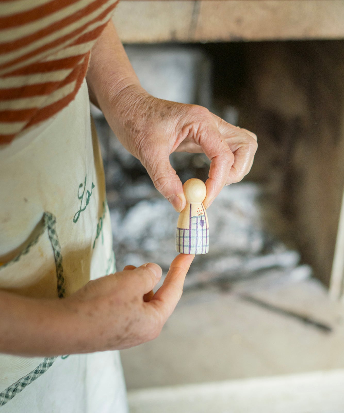 A person is holding a light coloured wooden Nin figure, with a pretty light blue and purple stripe apron painted onto it