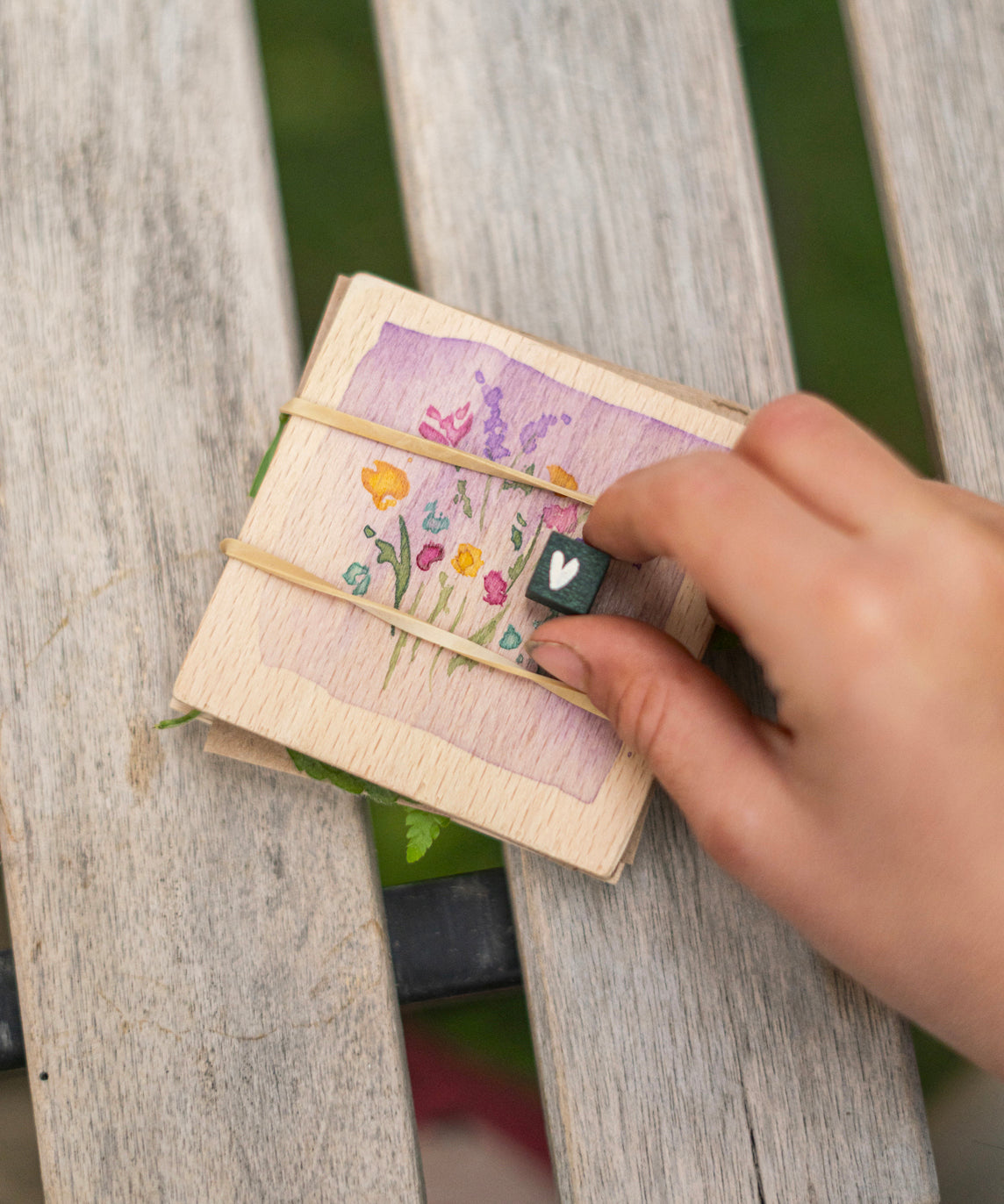 In the image is a wooden flower press activity from the Grapat Advent Calendar 2024. There is a lilac painted water-colour square on the top piece of wood, with delicately hand painted flowers, and is attached to another small square board by elastic bands. A child holds a small dark green cube with a white heart painted on the front. 