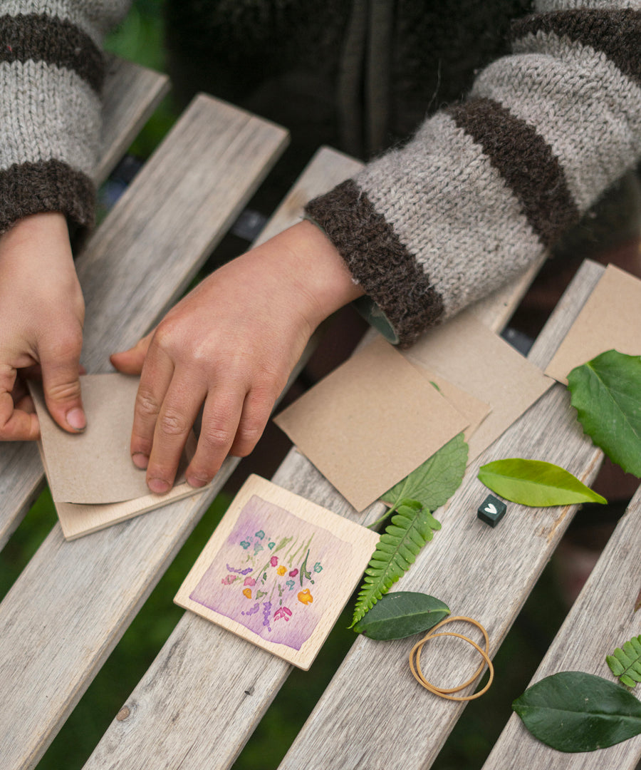 A child is playing with the wooden flower press in the Grapat Advent Calendar 2024. The child is placing light brown paper into the flower press, and will put green leaves in between the pieces of paper.