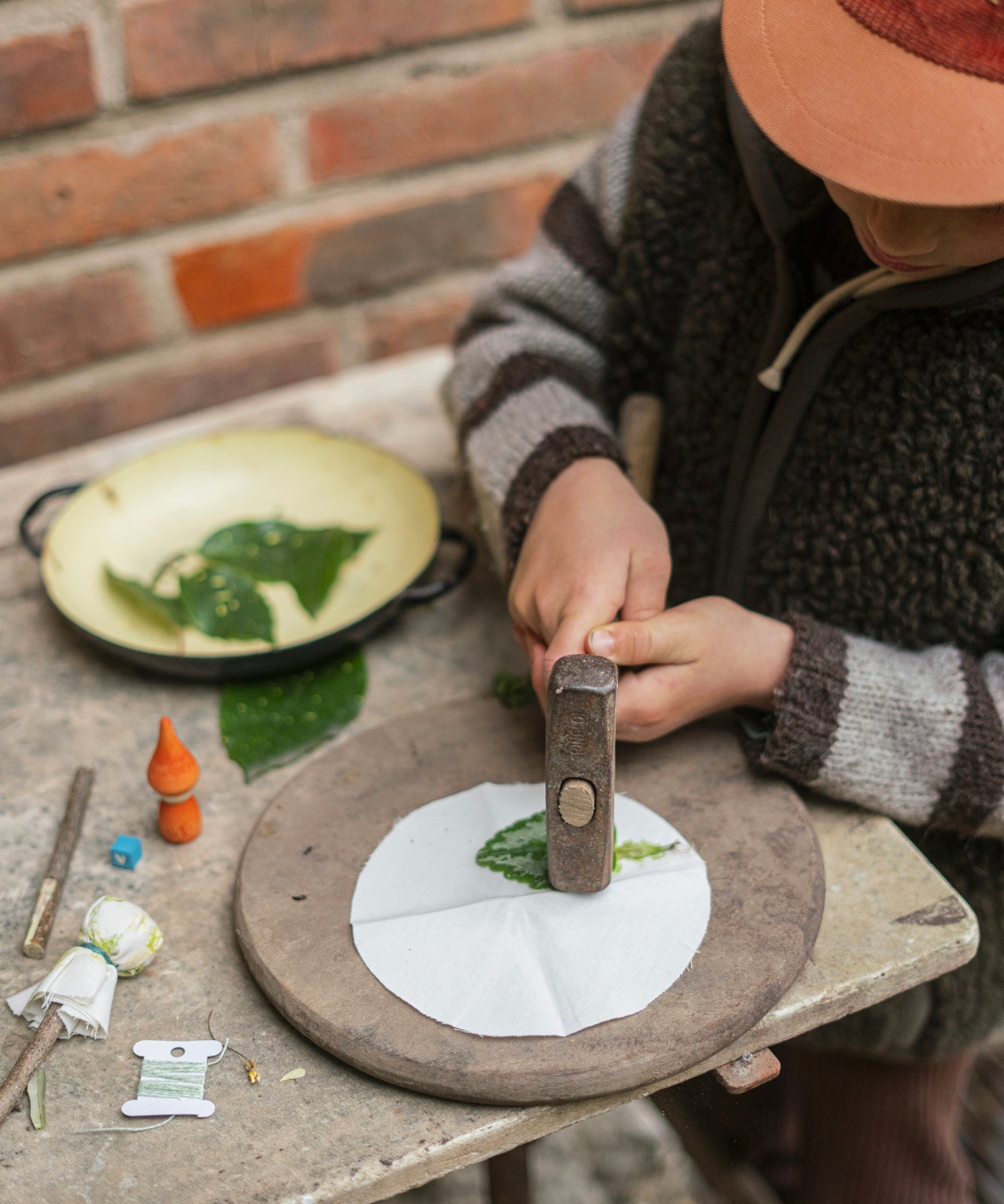 A child is hammering onto a leaf which is laid flat on a round, white piece of cloth. This image is showing the leaf pressing activity. Next to the child is a bowl of green leaves, are other items from the Grapat Advent Calendar 2024