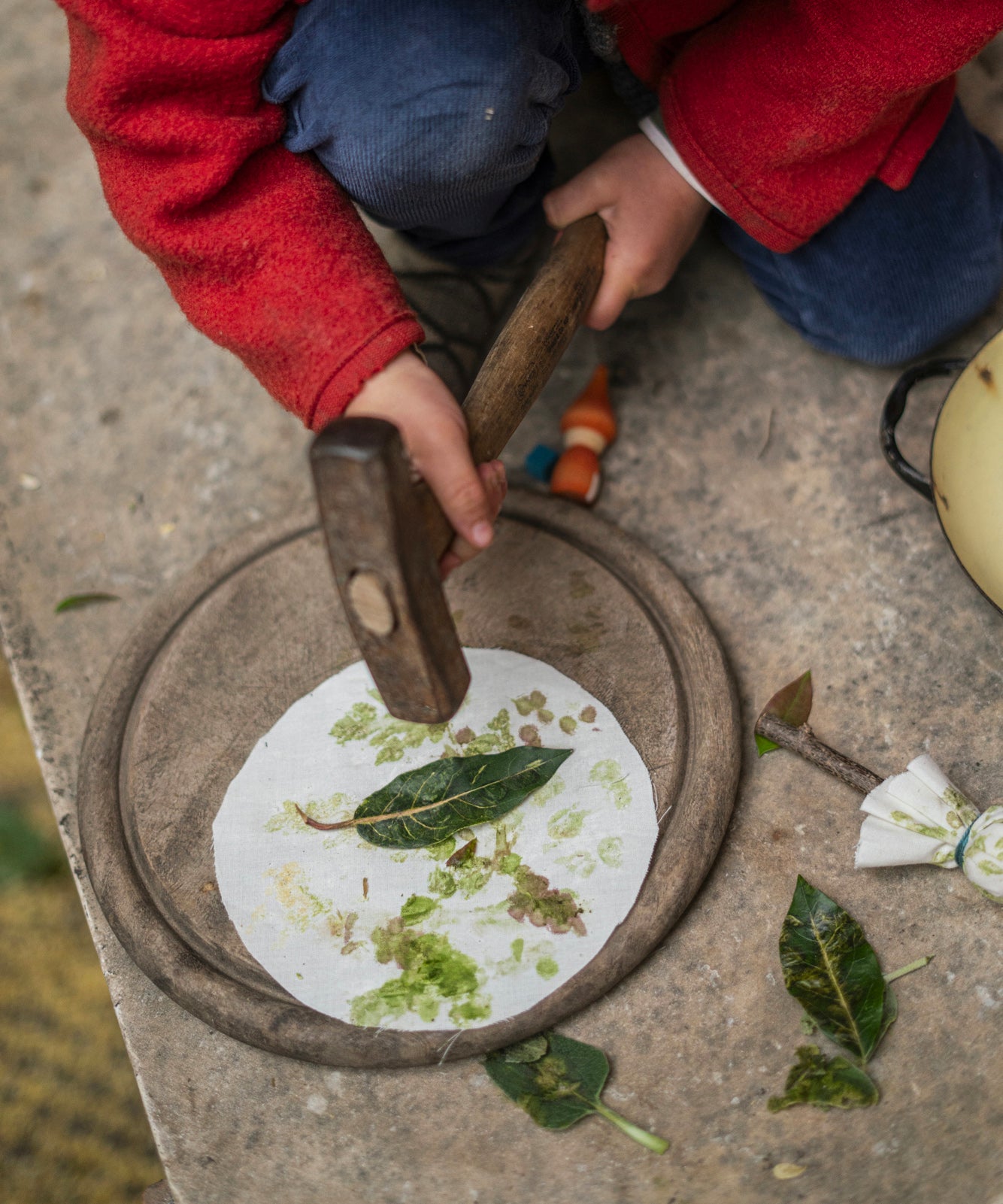 A closer view of the leaf art activity, with a child hammering a leaf on the top of a round white cloth to transfer the leaf colour onto the cloth