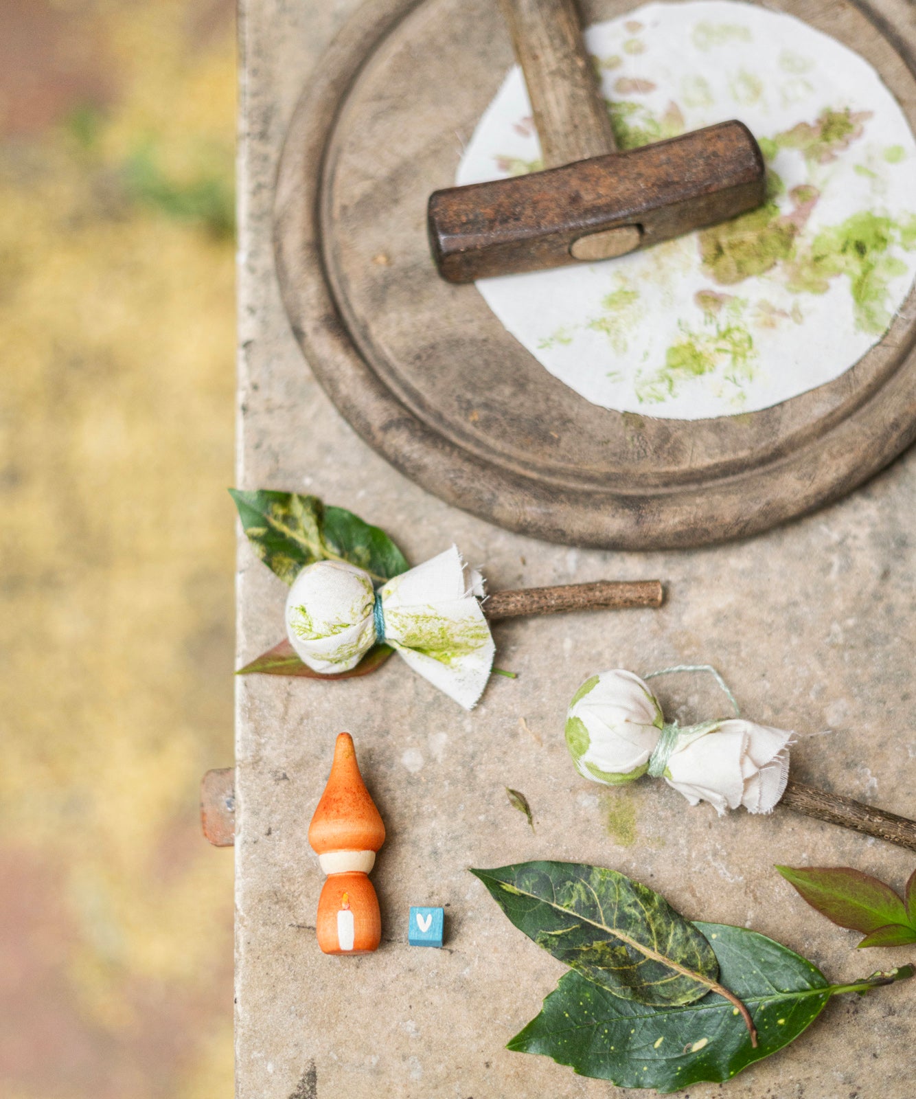 Various pieces of cloth set out on a concrete surface. There are two pieces of cloth wrapped onto wooden sticks, and a flat piece of circular cloth which has had leaves pressed into it to create amazing leaf art. In the image there is also a hammer, a small orange figure with a white candle painted on the front, and a small blue cube with what looks like a white letter V on the front