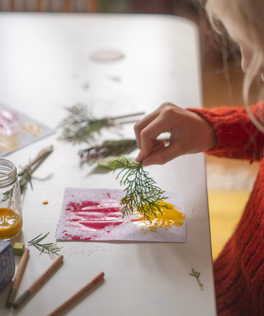 A child is creating art using red and yellow paint on a white piece of paper, and is using small green tree leaves and the paintbrush