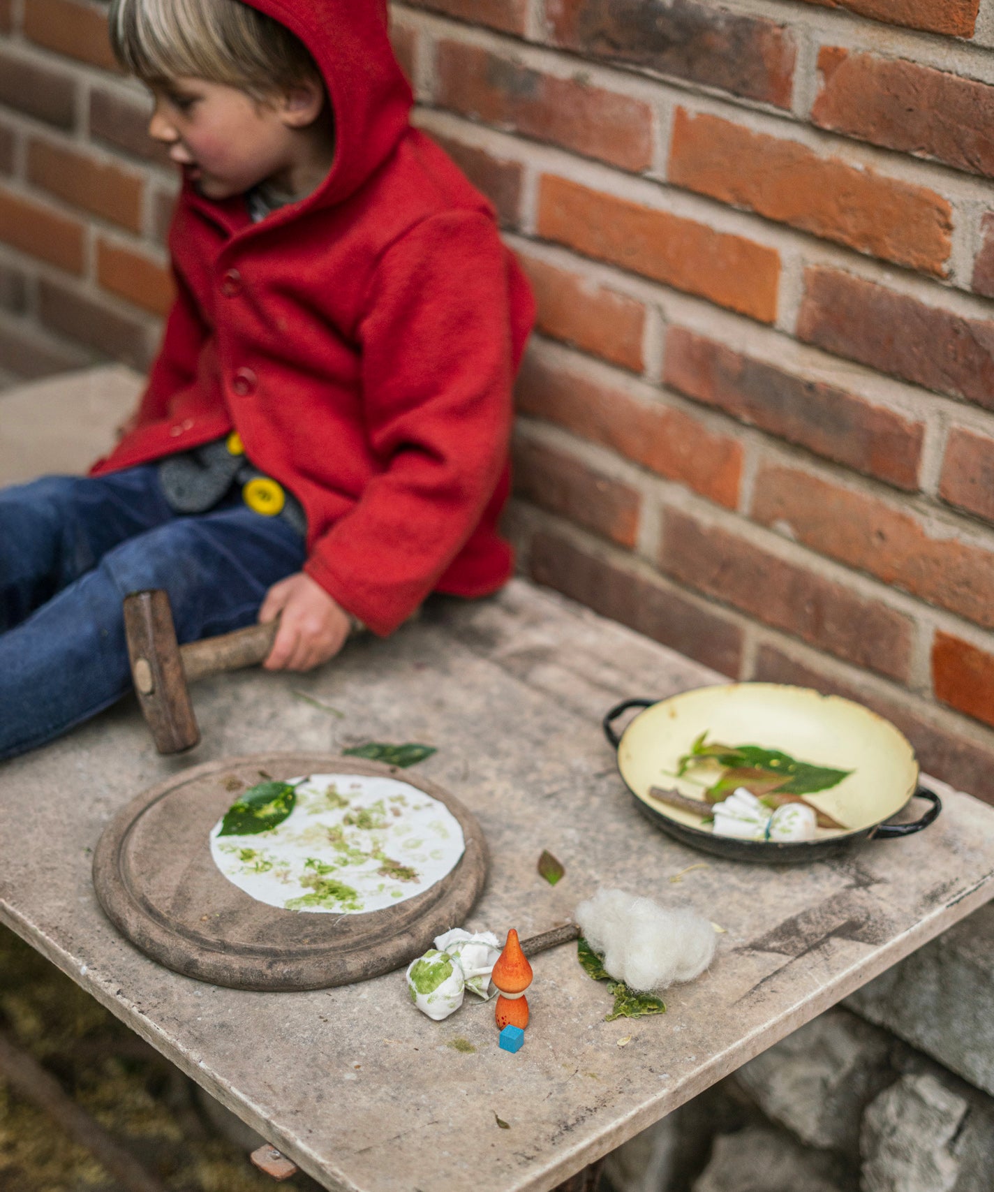 A child is sitting on a light coloured table, resting after creating lovely leaf art by hamming or pressing the leaves into white cloth so the colour of the leaf transfers onto the cloth