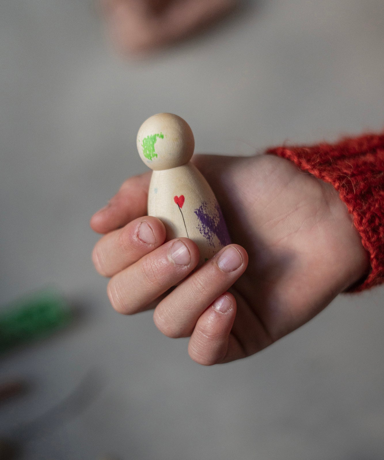 A child is holding a painted wooden Nin toy figure in their hand. The Nin is made from light wood with green and purple paint splotches, and a small red heart and a delicate black line coming from it, like a balloon. The Nin has been delicately painted by hand