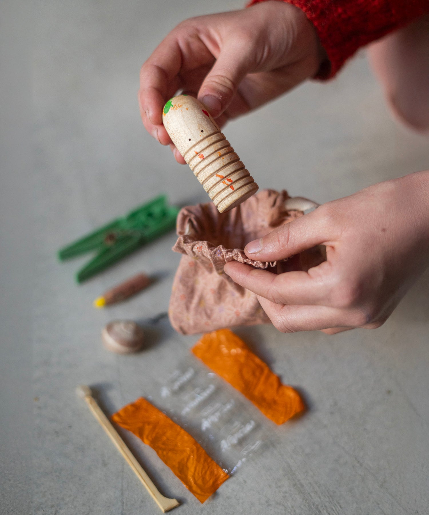 A child has a selection of loose play parts on the floor, and is putting a painted wooden figure into a peach coloured bag. The figure is light wood with orange and green painted splotches, two little black dots for eyes and cut in ridges to ad texture