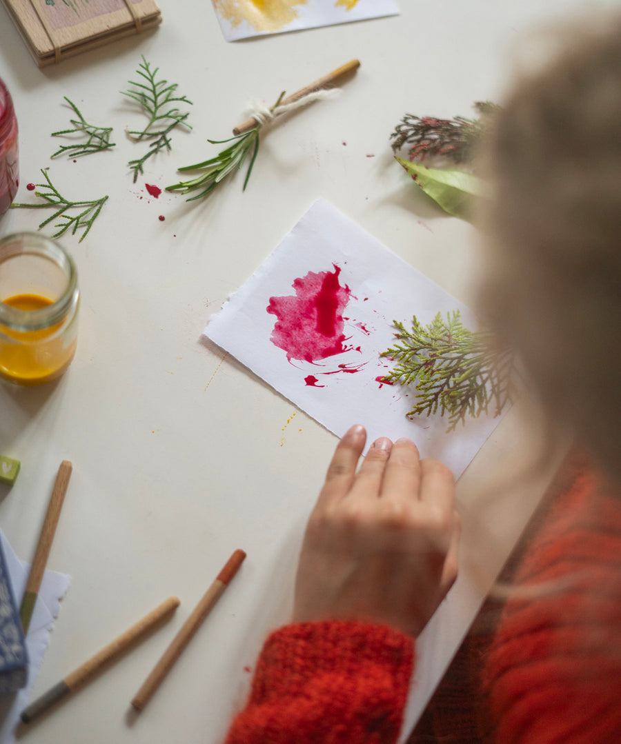 A child is creating a work of art, using a small green branch and a wooden stick as a paintbrush and using red paint on a white piece of paper