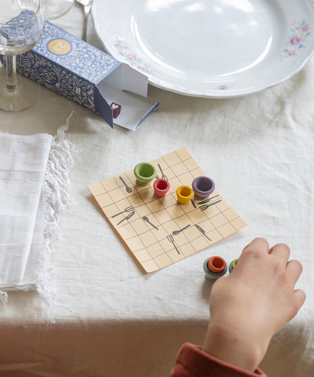 A small square of paper with lines in a grid pattern has little drawn knives, forks and spoons set out like a dinner placement. On top of the piece of paper are small, colourful wooden bowls which a child is arranging. On the table is a white cloth, a plate, a small glass and the 23rd box from the Grapat Advent Calendar 2024