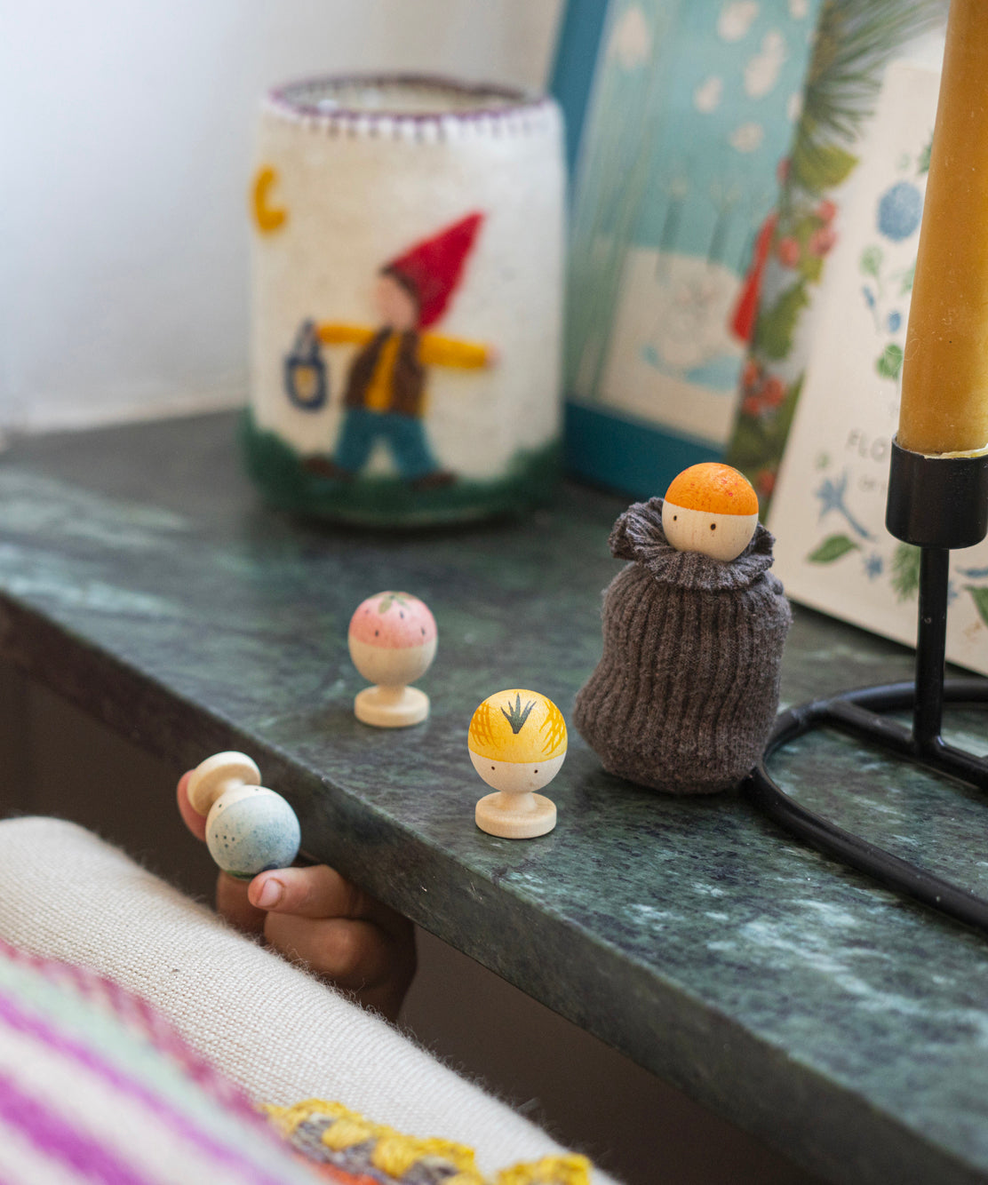 The colourful wooden fruit and vegetable figures are stood on a green/grey shelf with the blue one being held between a persons fingers and the orange figure in a sock, like a sock puppet