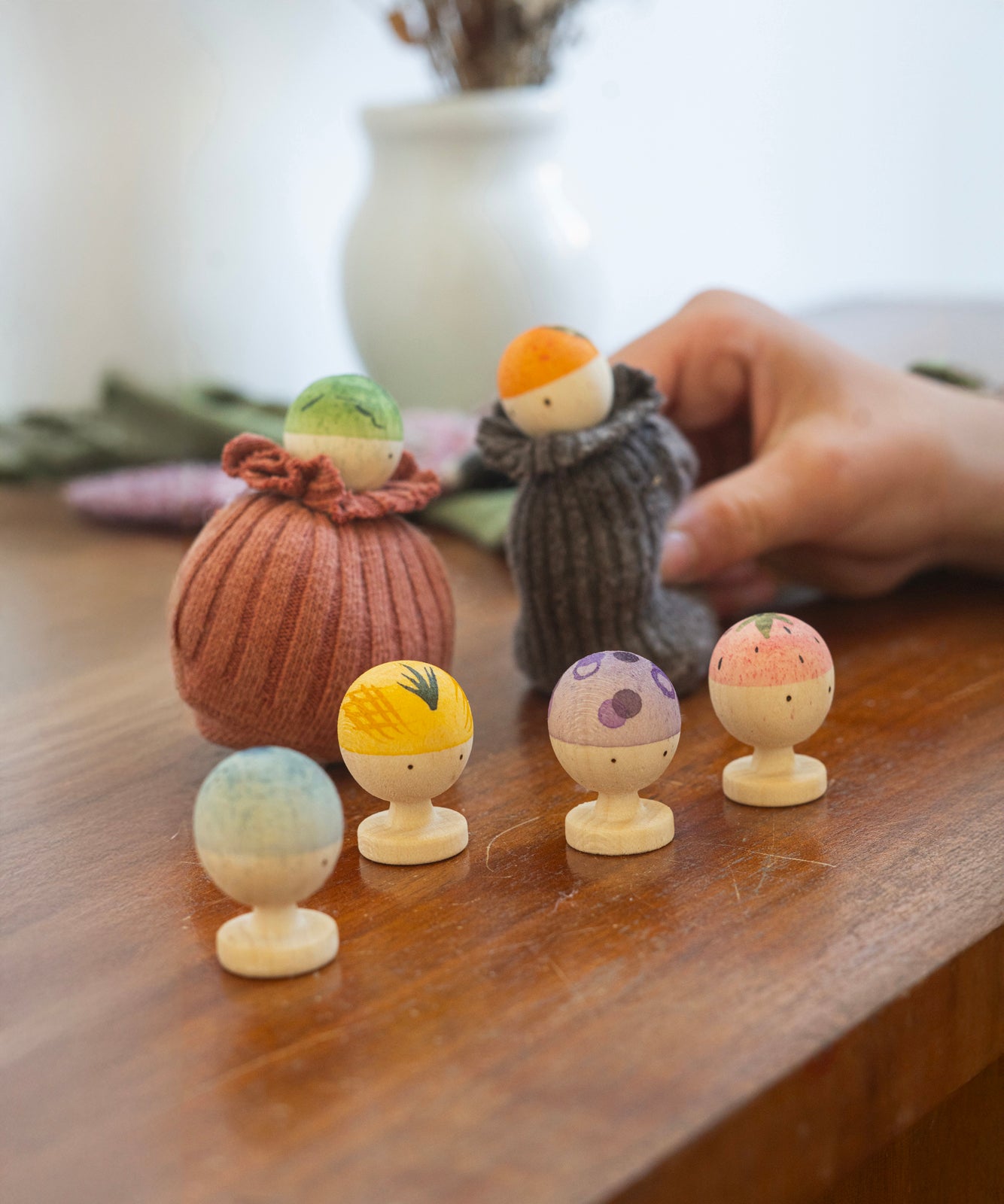 Six little wooden figures representing different fruits and vegetables. There is a green, orange, blue, yellow, purple and pink/red figure. With the green and orange figure inside a sock like a sock puppet