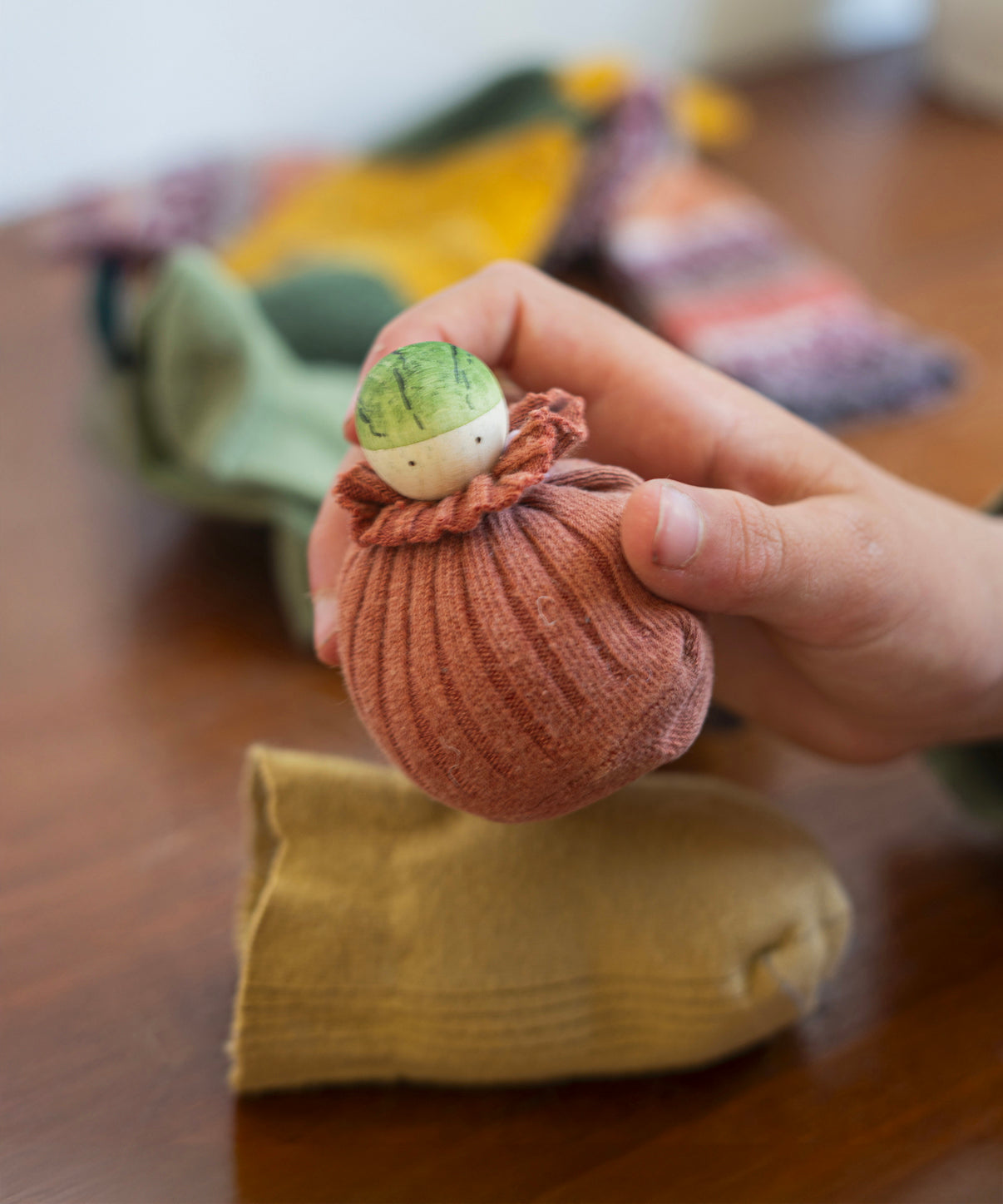 A child is holding what looks like a little wooden Brussels sprout figure, which is inside a sock like a sock puppet. The top of the little figure is various shades of green with two black dots as eyes. The figure has been carefully painted by hand., and blurred, colourful socks are laid out in the background