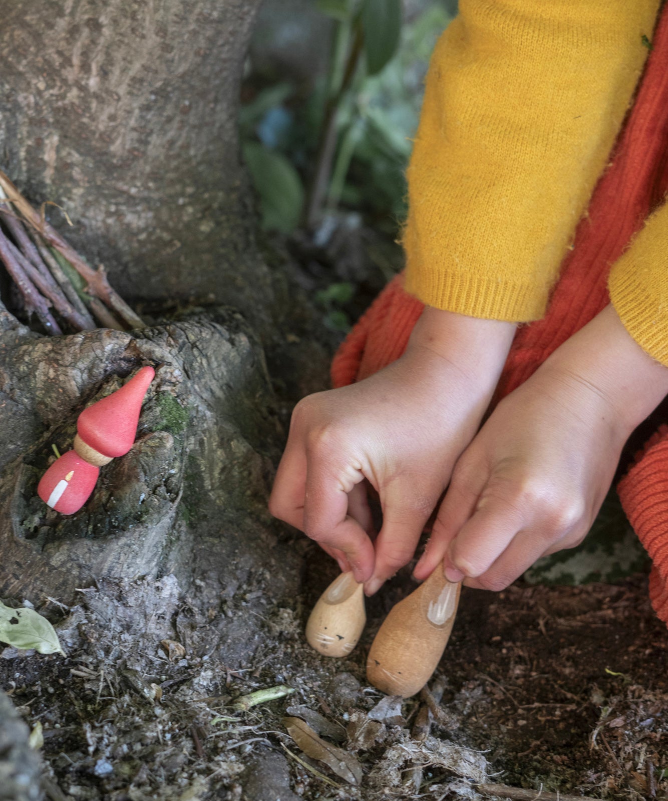 A closer look at the wooden rabbit figures being held by a child, and another wooden figure in red with a white candle painted on the front. The child is playing by a wooden tree trunk in a forest