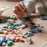 Child picking up a yellow wooden piece from the Grapat Mis Match & Bloom set, surrounded by scattered colourful loose parts on a terracotta-tiled floor. A woven basket filled with additional pieces sits nearby
