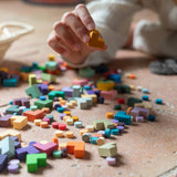 Child picking up a yellow wooden piece from the Grapat Mis Match & Bloom set, surrounded by scattered colourful loose parts on a terracotta-tiled floor. A woven basket filled with additional pieces sits nearby
