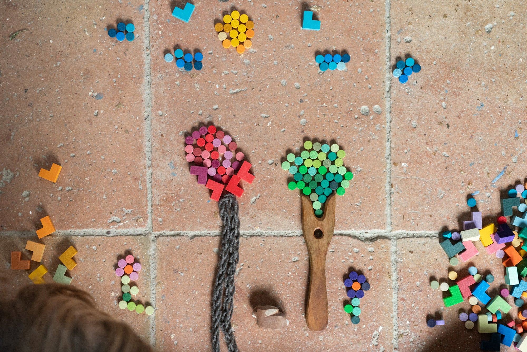 Creative play with the Grapat Mis Match & Bloom set, forming trees and flowers using colourful wooden loose parts. A wooden fork and braided yarn create tree trunks on a terracotta-tiled floor, with scattered pieces nearby.