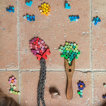 Creative play with the Grapat Mis Match & Bloom set, forming trees and flowers using colourful wooden loose parts. A wooden fork and braided yarn create tree trunks on a terracotta-tiled floor, with scattered pieces nearby.