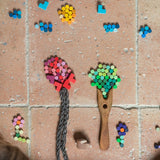 Creative play with the Grapat Mis Match & Bloom set, forming trees and flowers using colourful wooden loose parts. A wooden fork and braided yarn create tree trunks on a terracotta-tiled floor, with scattered pieces nearby.