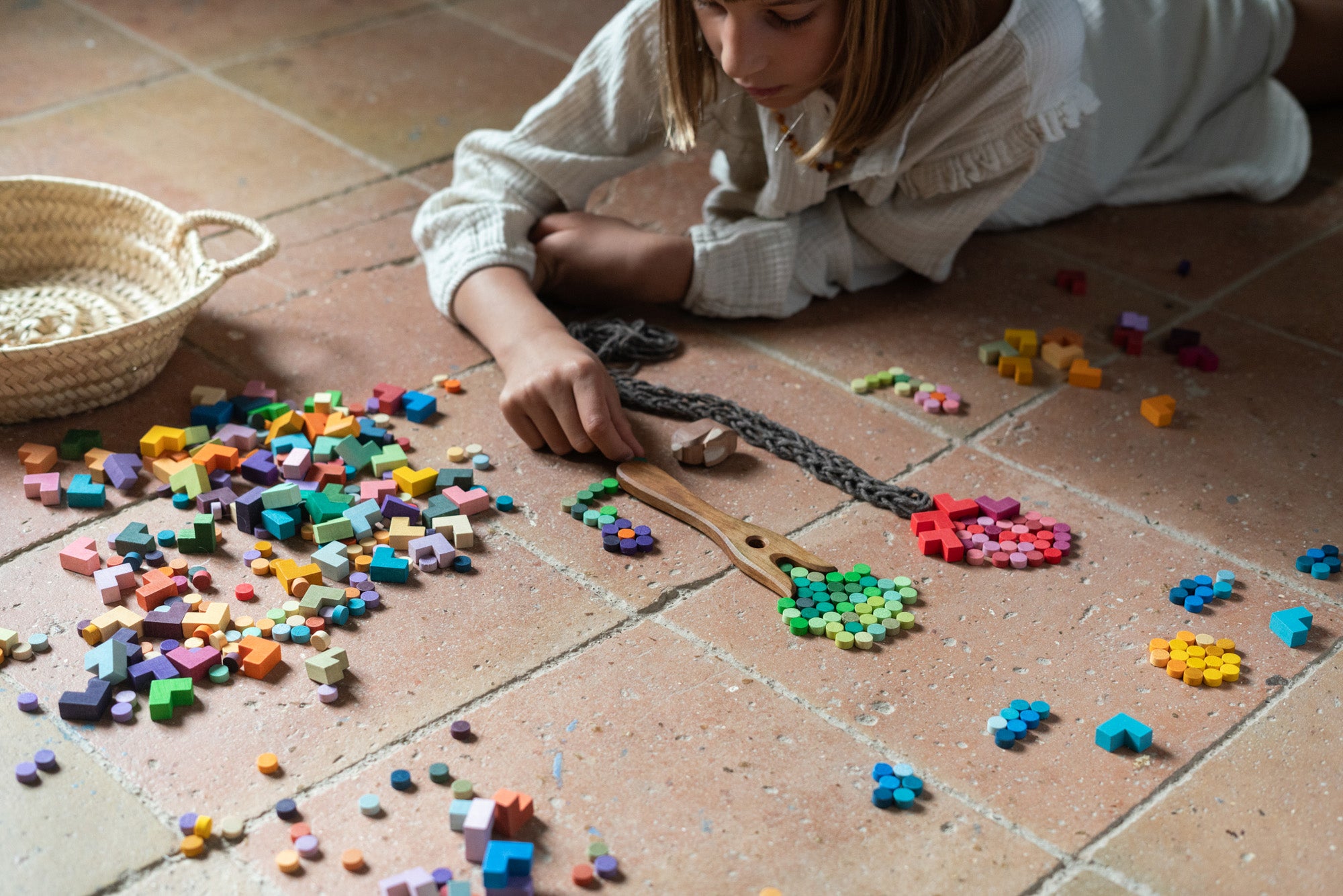 Child playing with the Grapat Mis Match & Bloom set, forming trees and flowers using colourful wooden loose parts. A wooden fork and braided yarn create tree trunks on a terracotta-tiled floor, with scattered pieces nearby.