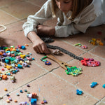 Child playing with the Grapat Mis Match & Bloom set, forming trees and flowers using colourful wooden loose parts. A wooden fork and braided yarn create tree trunks on a terracotta-tiled floor, with scattered pieces nearby.