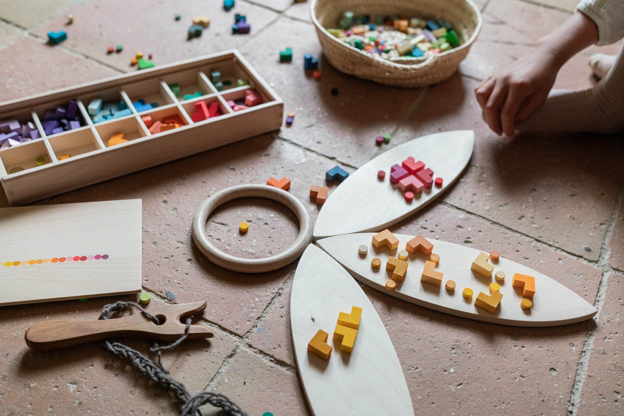 A child playing with the Grapat wooden bloom mis and match wooden block set. Featuring wooden Grapat petals a wooden fork and braided yarn and a woven basket on a terracotta floor