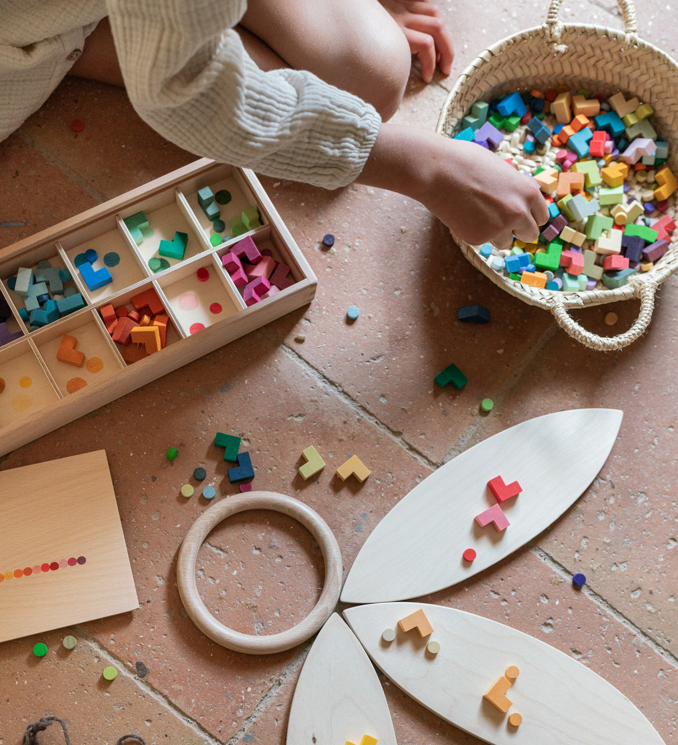 Child playing with the Grapat Mis Match & Bloom set, featuring a wooden sorting box, colourful geometric pieces, Grapat Petals, and a wooden ring on a terracotta-tiled floor.