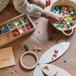 Child playing with the Grapat Mis Match & Bloom set, featuring a wooden sorting box, colourful geometric pieces, Grapat Petals, and a wooden ring on a terracotta-tiled floor.