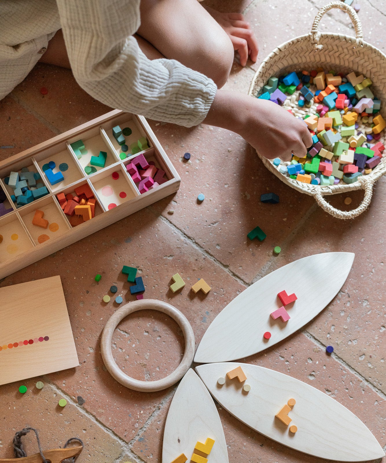 Child playing with the Grapat Mis Match & Bloom set, featuring a wooden sorting box, colourful geometric pieces, Grapat Petals, and a wooden ring on a terracotta-tiled floor.