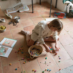 Child playing with the Grapat Mis Match & Bloom set featuring colourful geometric pieces and a woven basket with scattered colourful geometric pieces on a terracotta floor.