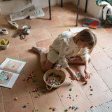 Child playing with the Grapat Mis Match & Bloom set featuring colourful geometric pieces and a woven basket with scattered colourful geometric pieces on a terracotta floor.