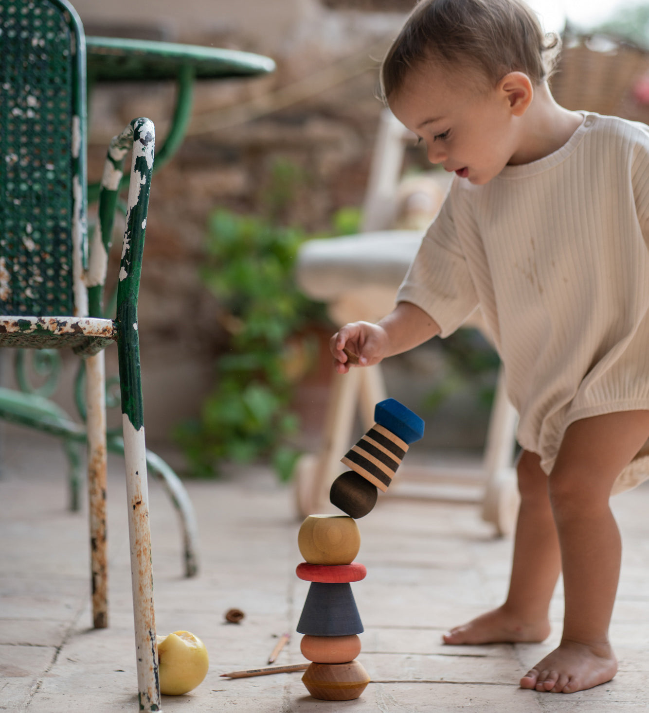 Child playing with the Grapat wooden serendipity stacking set outside on the floor next to a garden chair . Showing the different handcrafted tactile pieces stacked on the stone floor. Features a brown, light pink, blue yellow orange and striped pieces. 