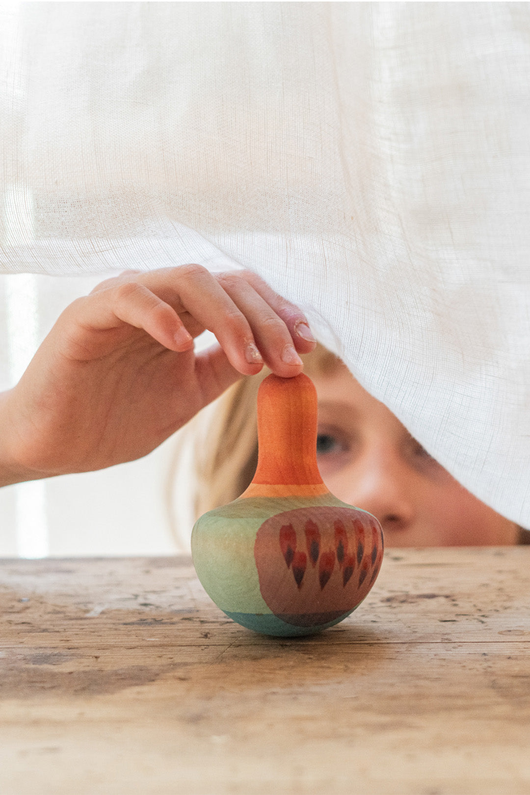 A child carefully lifts a Grapat Wooden Wobbly Bird in Mellow Orange, partially hidden behind a flowing white curtain. The moment captures the magic of discovery and mindful play, reflecting the essence of Grapat's All the Nothing collection—available now at Babipur.