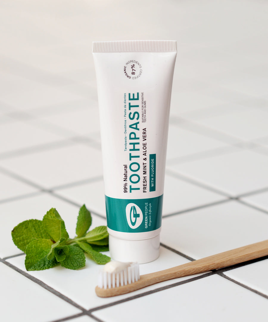 The Green People Fresh Mint & Aloe Vera Toothpaste with Fluoride is stood on a white tiled floor, with a bamboo tooth brush in front and a sprig of fresh mint leaves to the side