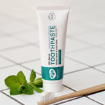 The Green People Fresh Mint & Aloe Vera Toothpaste with Fluoride is stood on a white tiled floor, with a bamboo tooth brush in front and a sprig of fresh mint leaves to the side