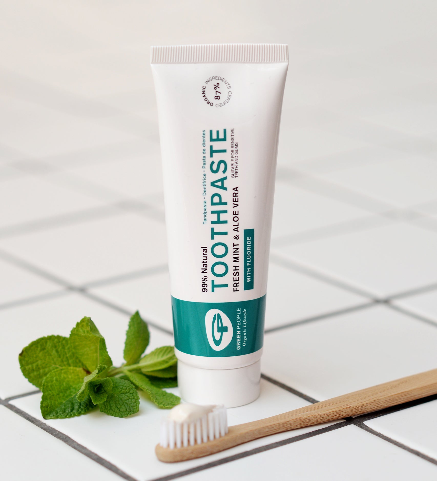 The Green People Fresh Mint & Aloe Vera Toothpaste with Fluoride is stood on a white tiled floor, with a bamboo tooth brush in front and a sprig of fresh mint leaves to the side