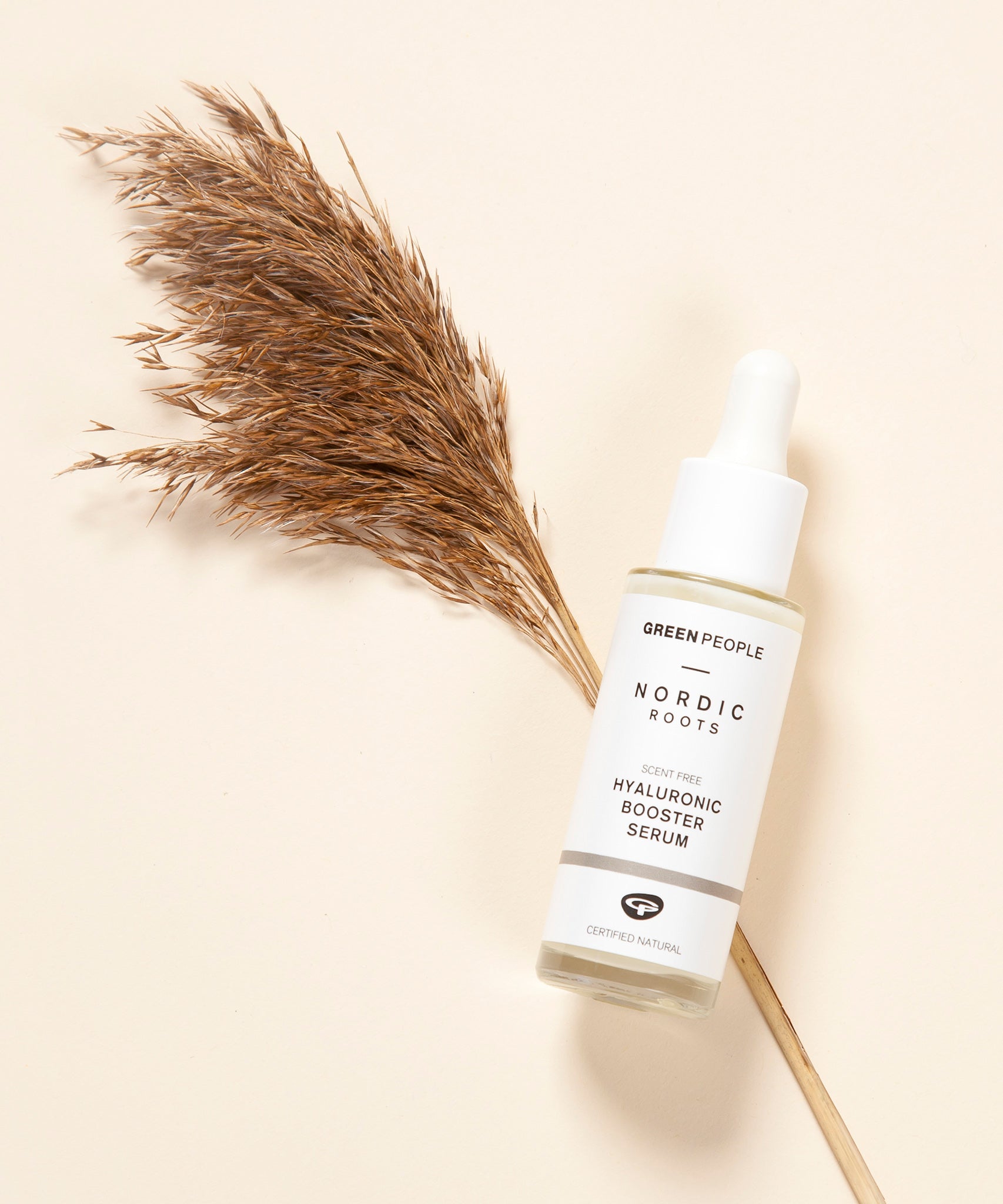 The Green People Nordic Roots Hyaluronic Booster Serum on a light peach background with dried grass underneath