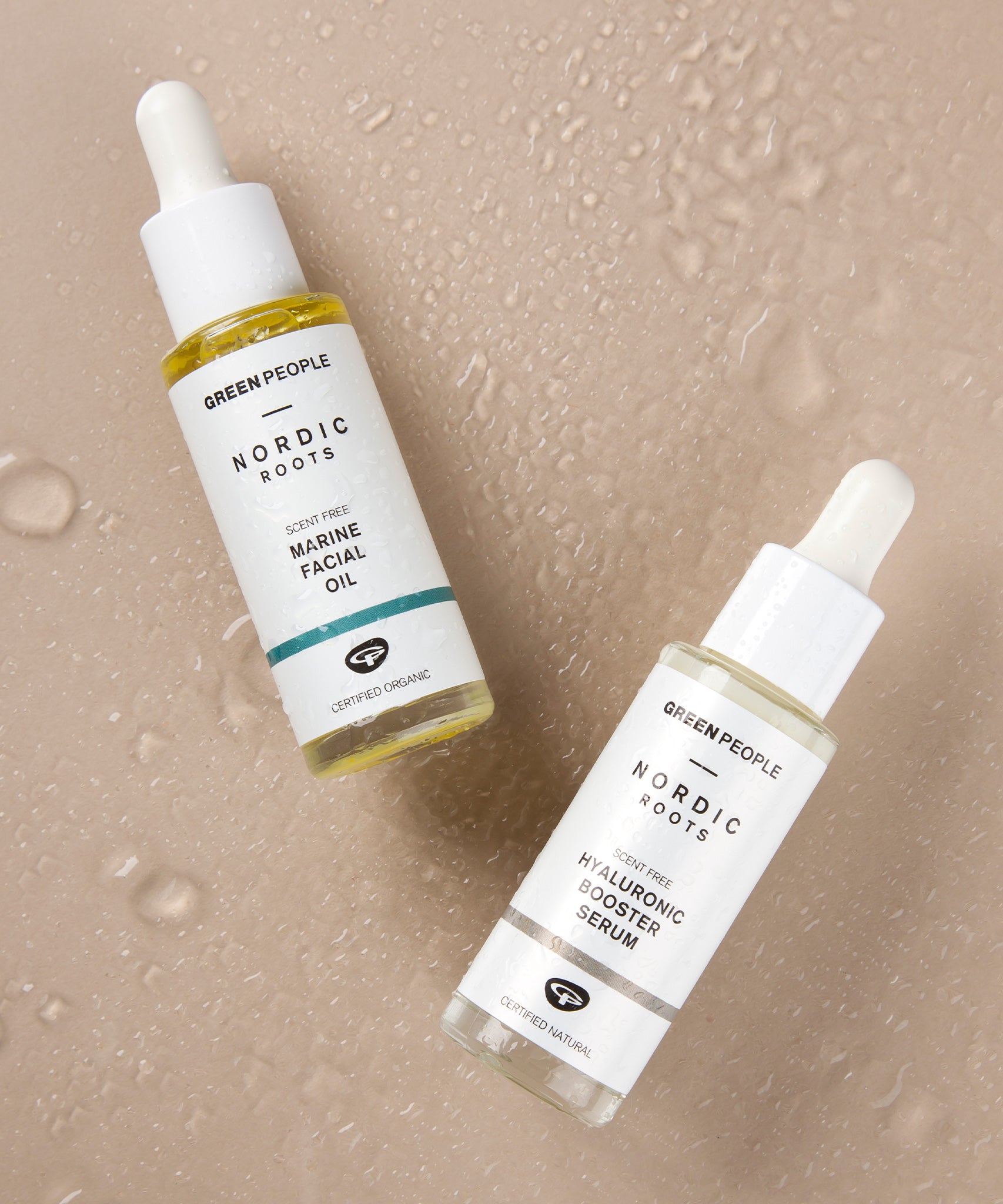 The Green People Nordic Roots Hyaluronic Booster Serum and Nordic Roots Marine Facial Oil laid on a peach coloured background with water droplets, to represent the hydrating nature of the products