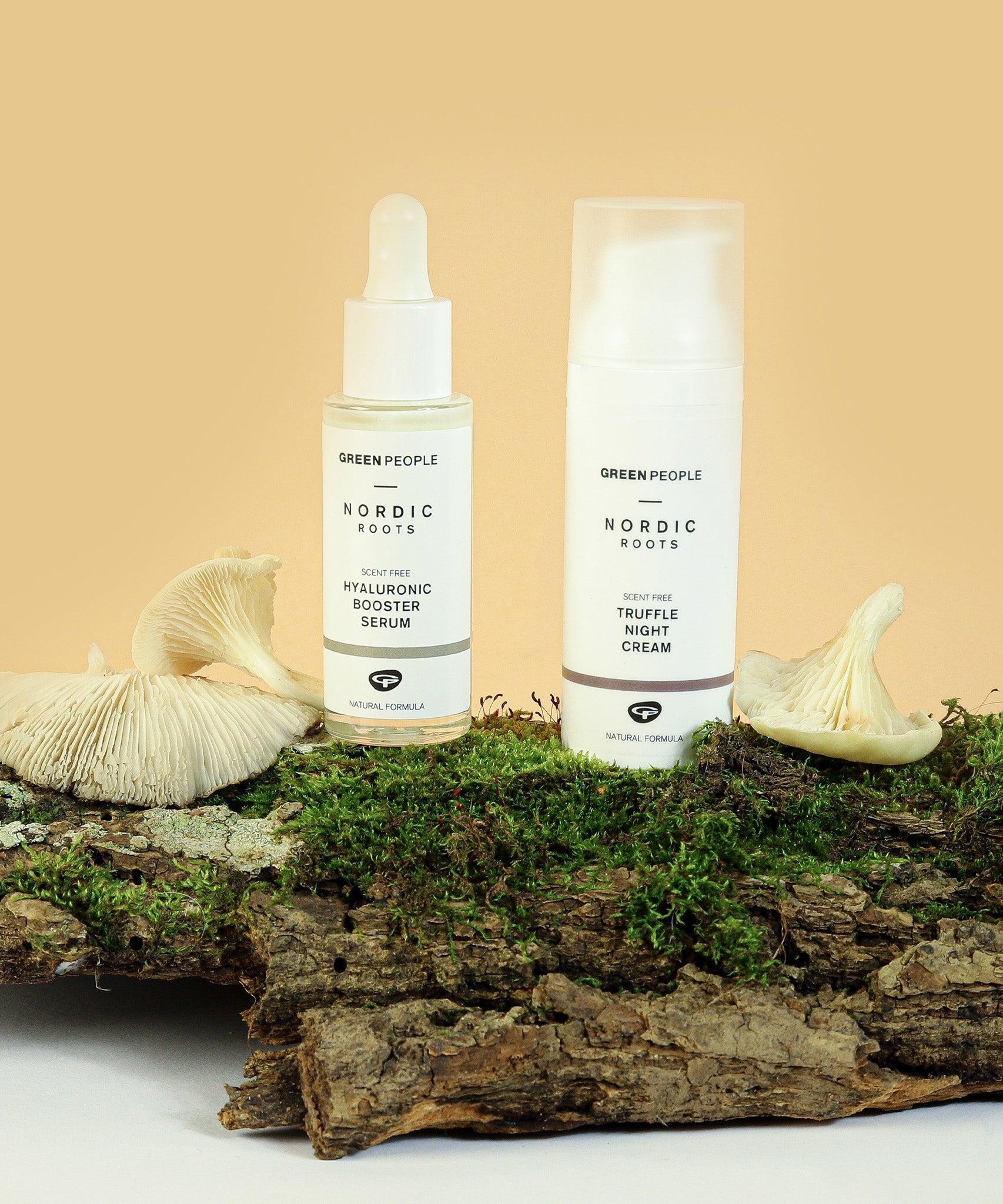 The Green People Nordic Roots Hyaluronic Booster Serum and Truffle Night Cream are placed upright on a wooden log with moss and chanterelle mushrooms, to represent the natural ingredients used in the Nordic Roots range