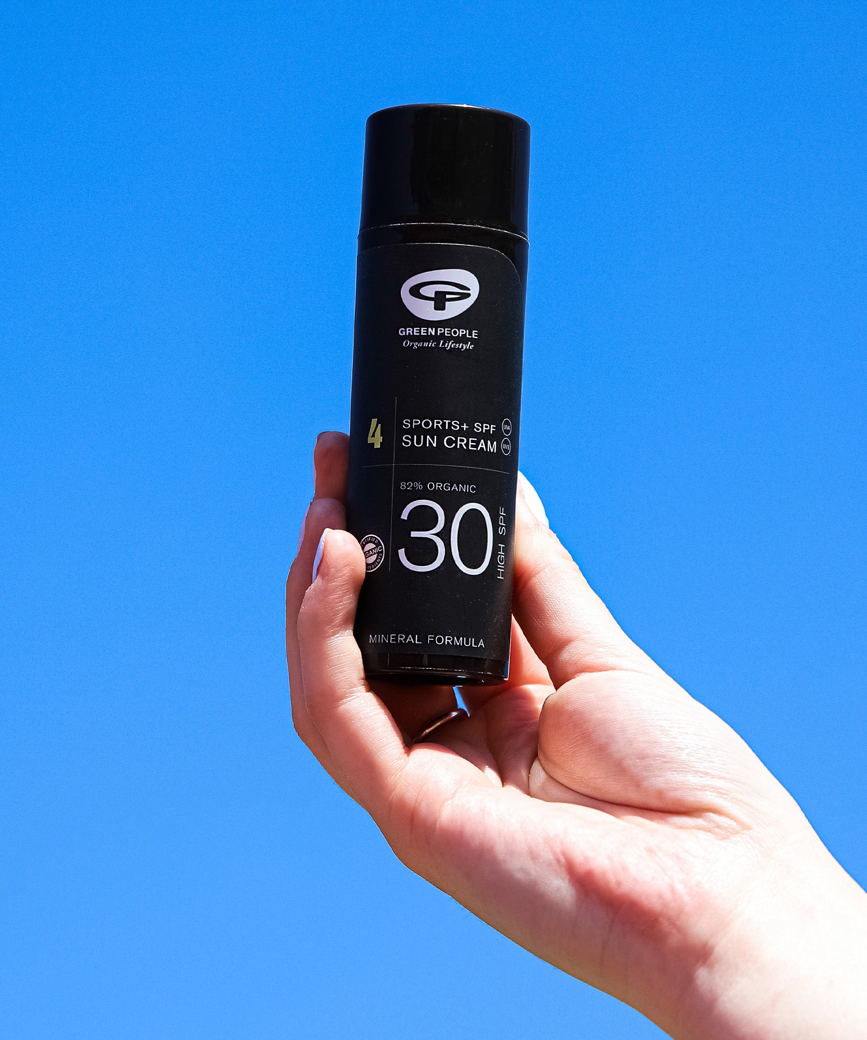 A person holding the Green People Sports SPF30 Mineral Facial Sun Cream 50ml in their hand, with a blue sky in the background