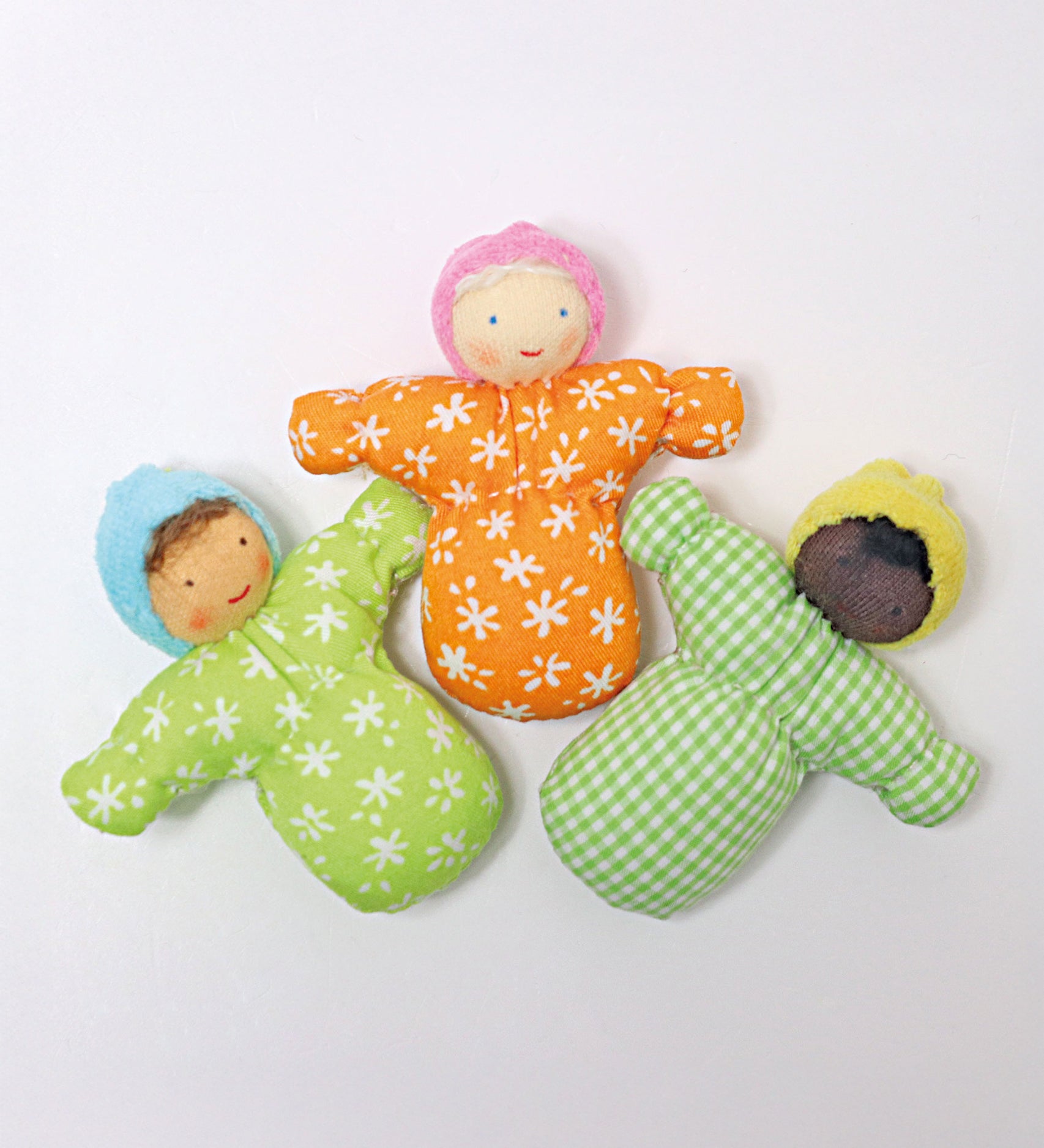 Three Grimm's Waldorf Baby doll toys laid out next to each other. The left doll Baby Leo has a light green body with white flower print, a light blue hat, and light brown hair. The middle doll Baby Mia, has an orange body with white flower print, a light pink hat and white hair, and the left doll Baby Sam has a green check print body, a yellow hat and black hair.