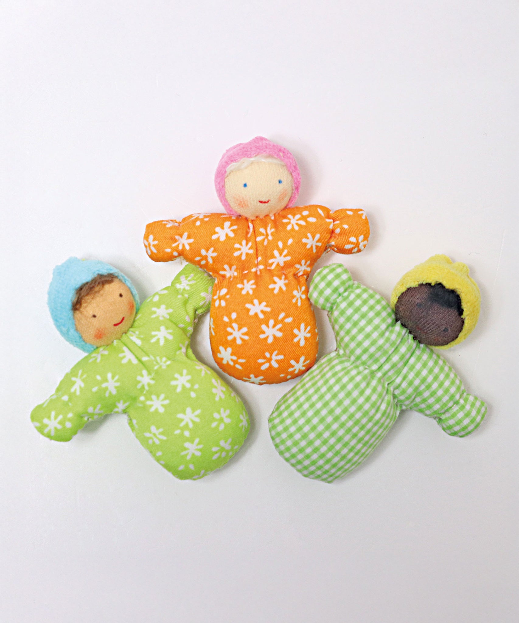 Three Grimm's Waldorf Baby doll toys laid out next to each other. The left doll Baby Leo has a light green body with white flower print, a light blue hat, and light brown hair. The middle doll Baby Mia, has an orange body with white flower print, a light pink hat and white hair, and the left doll Baby Sam has a green check print body, a yellow hat and black hair.