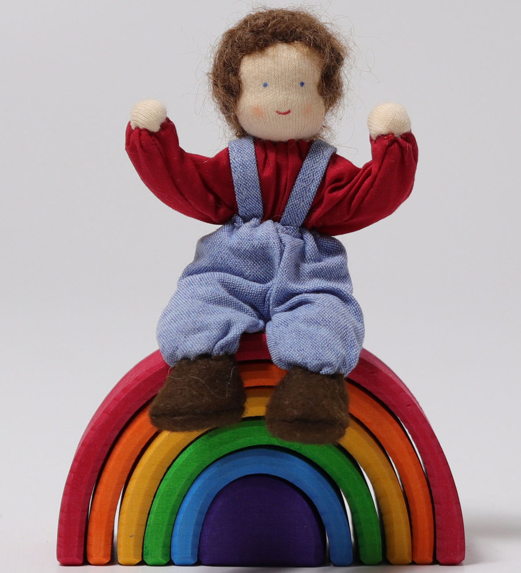 A closer look at Peter, sat on a Grimm's small wooden rainbow, with its arms bent upwards to show the dolls flexibility, on a light grey background. Peter is a handmade, Waldorf, fabric doll.