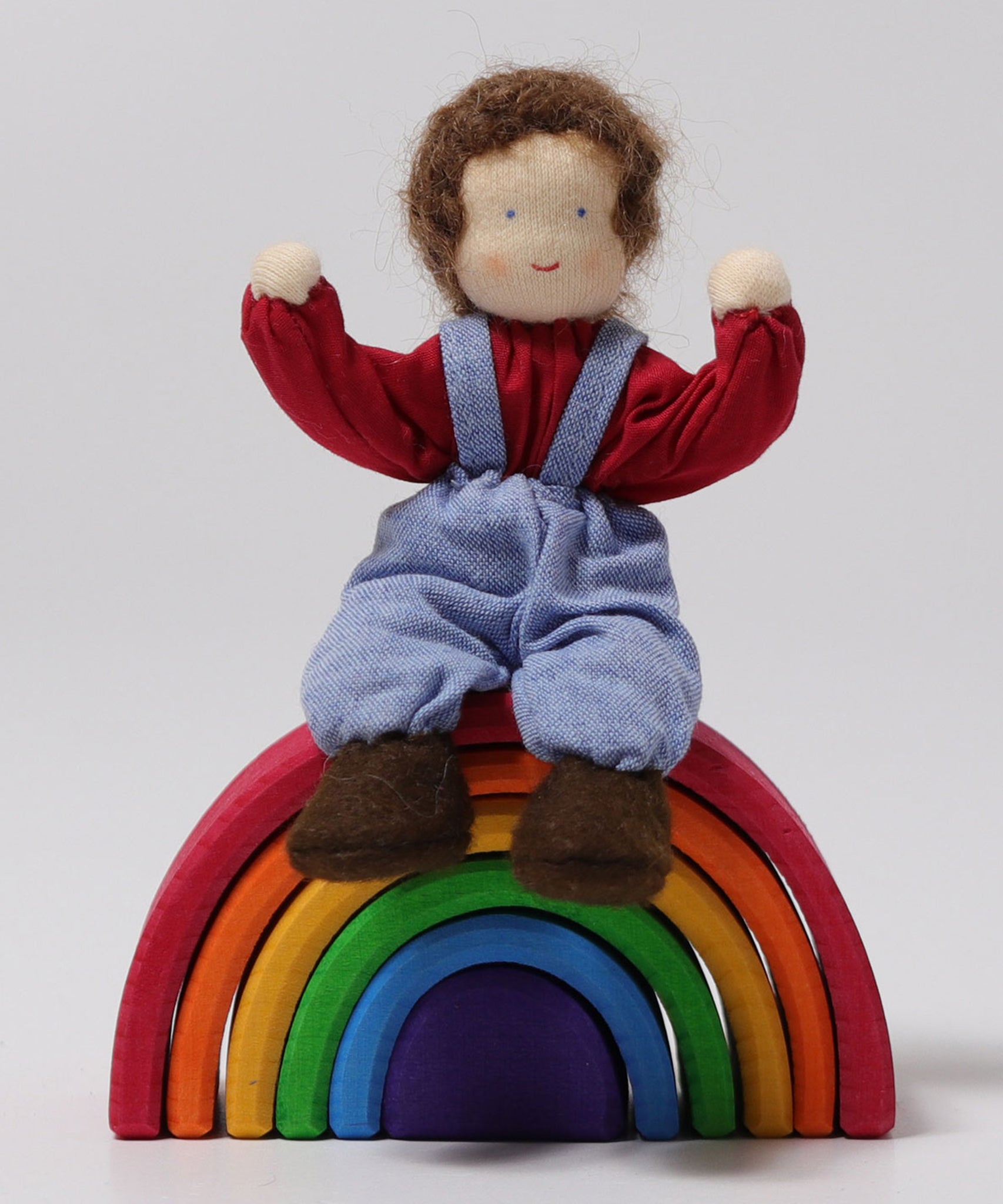 A closer look at Peter, sat on a Grimm's small wooden rainbow, with its arms bent upwards to show the dolls flexibility, on a light grey background. Peter is a handmade, Waldorf, fabric doll.