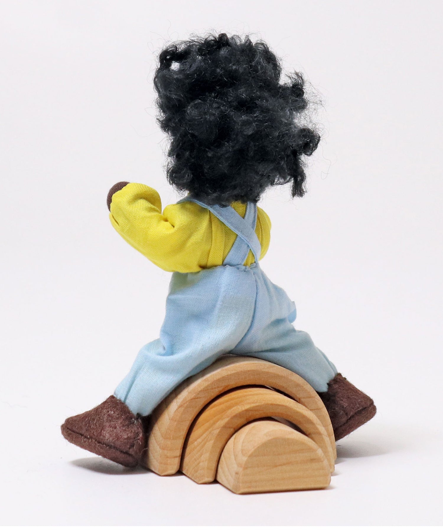 A view of Grimm's Handmade Doll - Max from the back. Max is climbing over a Grimm's small natural rainbow to show how flexible its arms and legs are. The image is on a light grey background