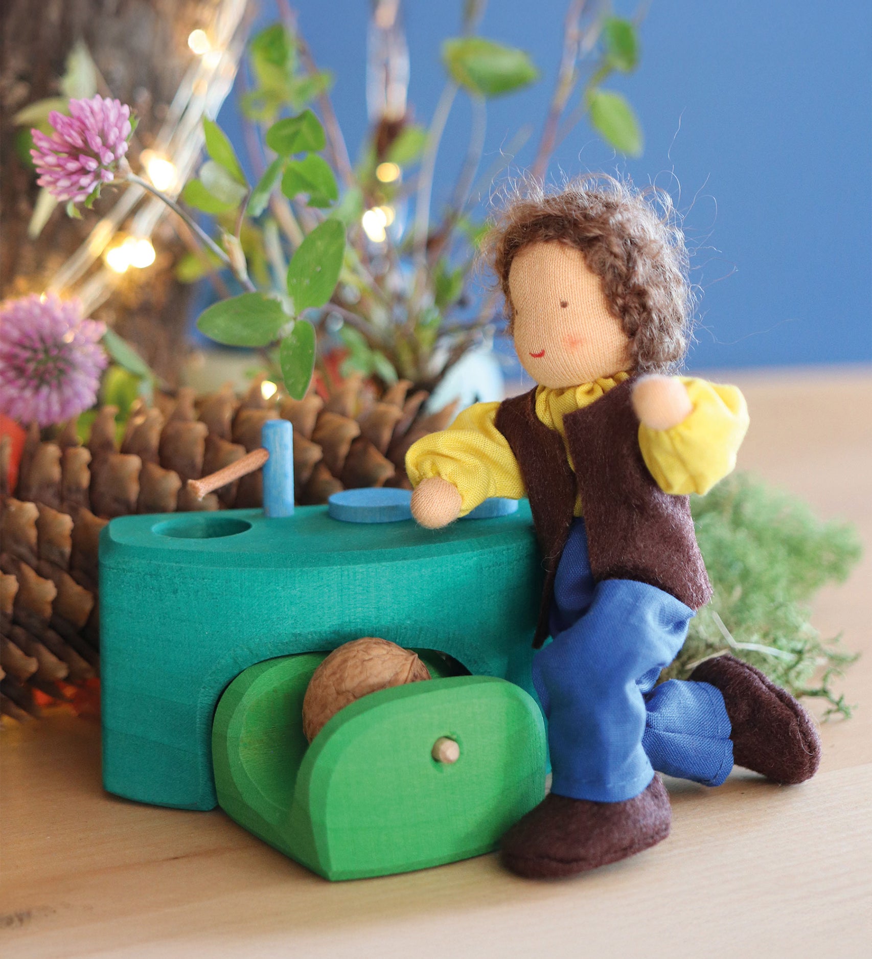 Grimm's Brown Haired Man Doll - Erle is bent down on its knees, next to a little green wooden kitchen. There are flowers and thin tree branches in the background.