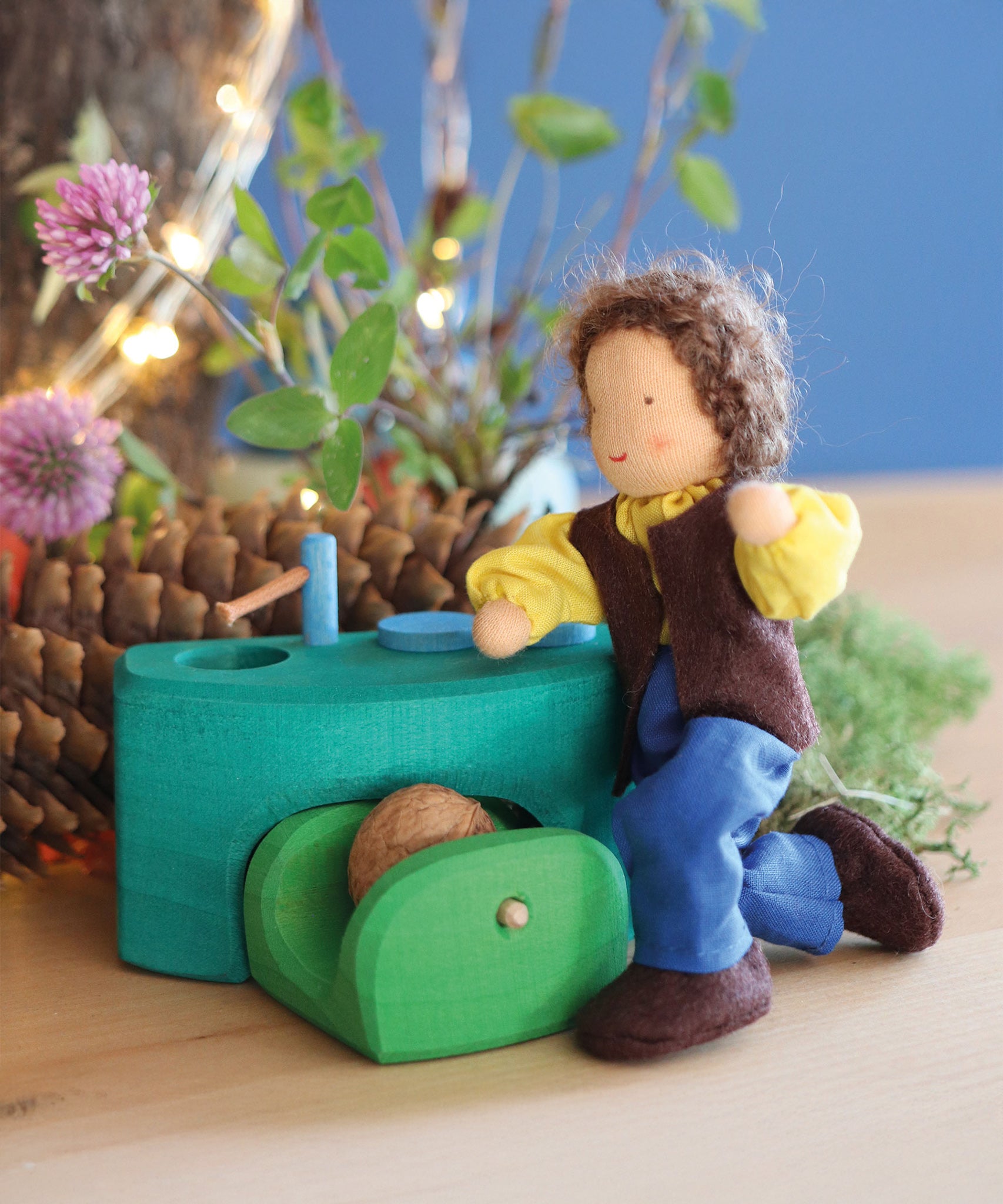 Grimm's Brown Haired Man Doll - Erle is bent down on its knees, next to a little green wooden kitchen. There are flowers and thin tree branches in the background.