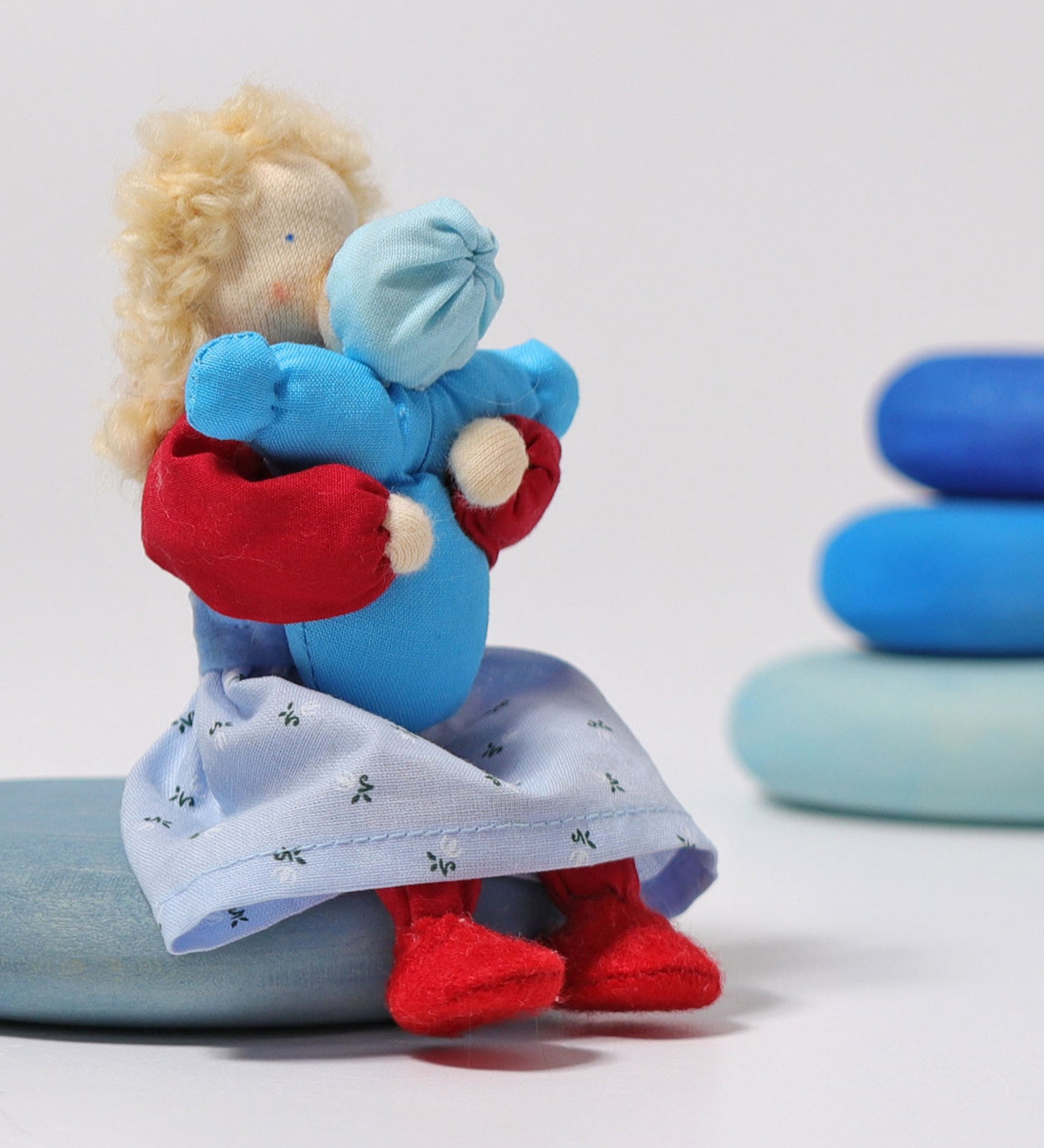 The Grimm's Blonde Woman Doll, is holding a small doll representing a child which is dressed in blue, and is sitting on a blue Grimm's Wooden river pebbles piece