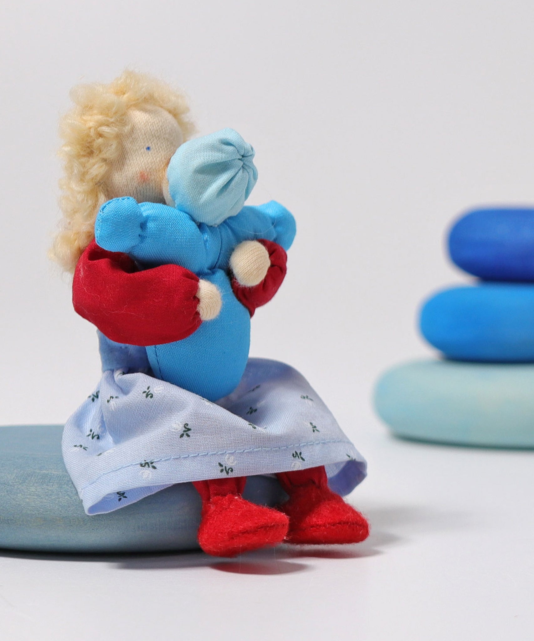 The Grimm's Blonde Woman Doll, is holding a small doll representing a child which is dressed in blue, and is sitting on a blue Grimm's Wooden river pebbles piece