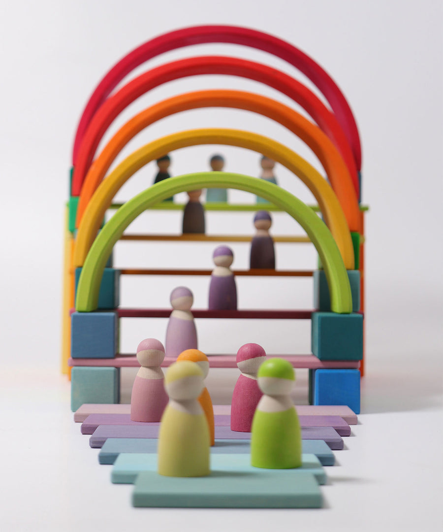 Grimm's 12 Pastel Friends stood on colourful pastel Grimm's blocks and rainbow arches. The image shows the Grimm's Pastel friends in the foreground and in the backgrund stood on Grimm's wooden pastel coloured slats, underneath the rainbow arches